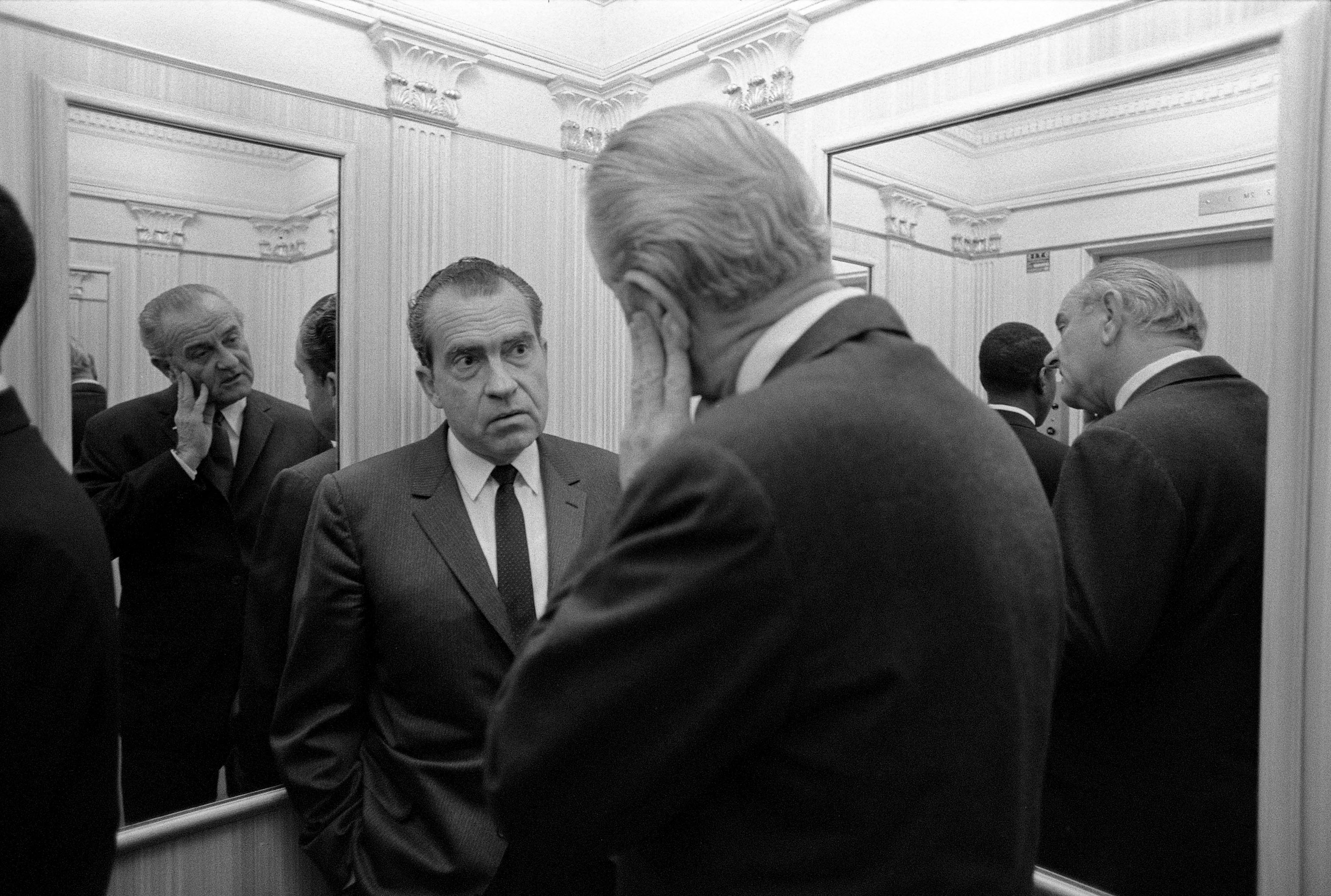 President Lyndon B. Johnson, foreground with his back to the camera, speaks to Richard Nixon in an elevator. Mirrors on either side reflect President Johnson's face.